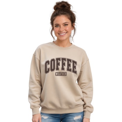 Woman wearing a sand crewneck sweatshirt with "COFFEE WEATHER" printed in dark brown varsity letters, paired with blue jeans against a white background.