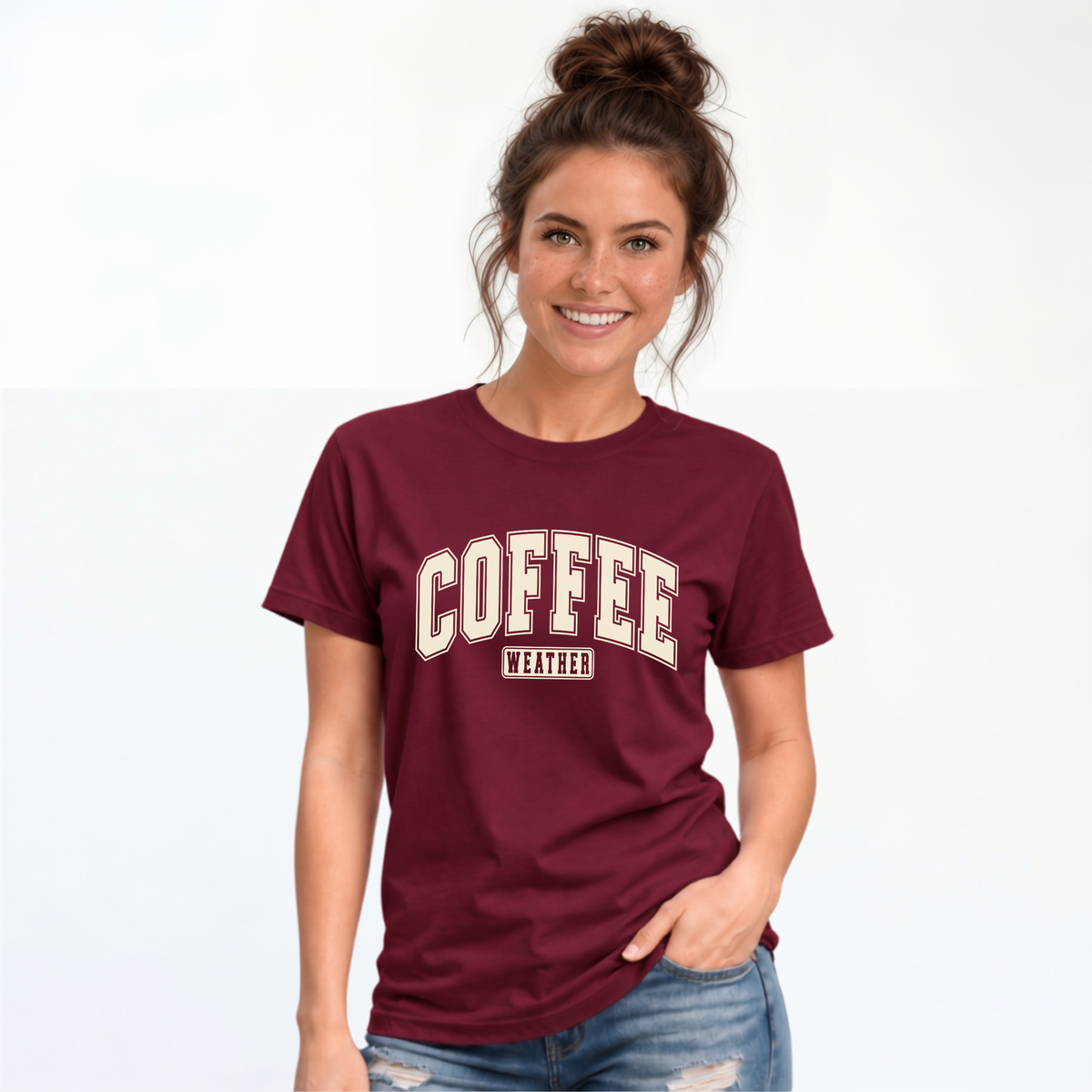 Woman modeling a maroon T-shirt with "COFFEE WEATHER" printed in cream and navy on the front, paired with distressed blue jeans against a white background.