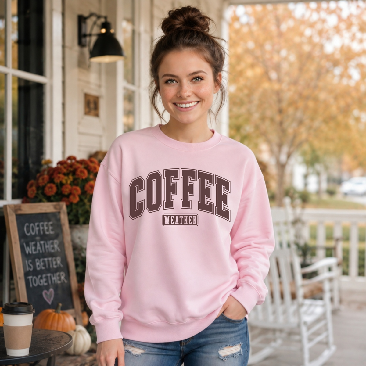 Woman standing on a porch wearing a light pink crewneck sweatshirt with "COFFEE WEATHER" printed in dark brown across the chest.