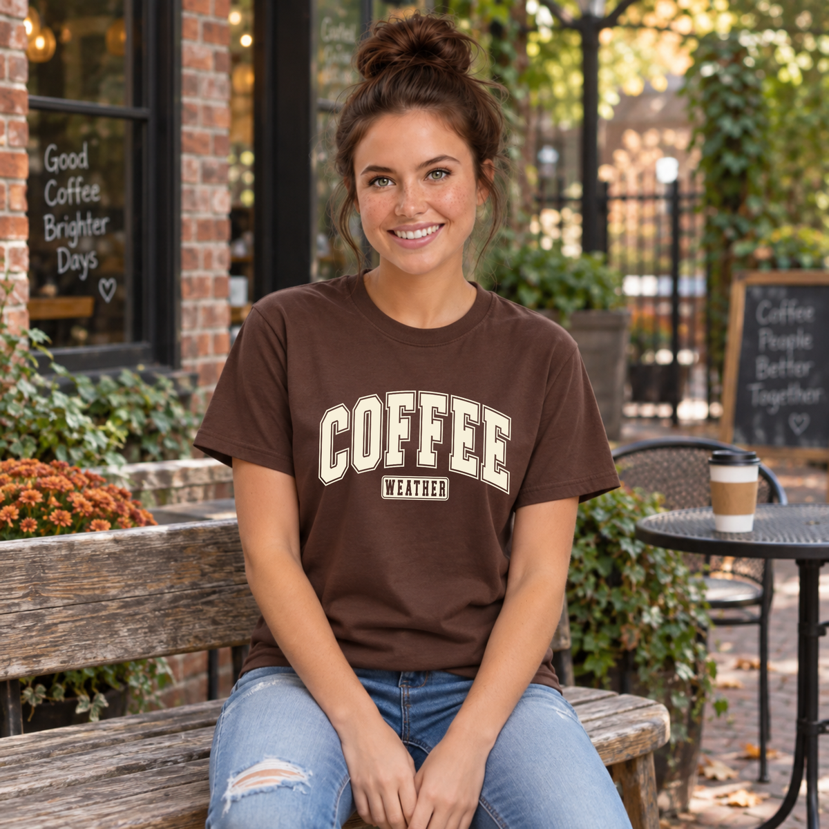 Woman modeling a brown T-shirt with "COFFEE WEATHER" printed in cream varsity lettering on the front against a white background.