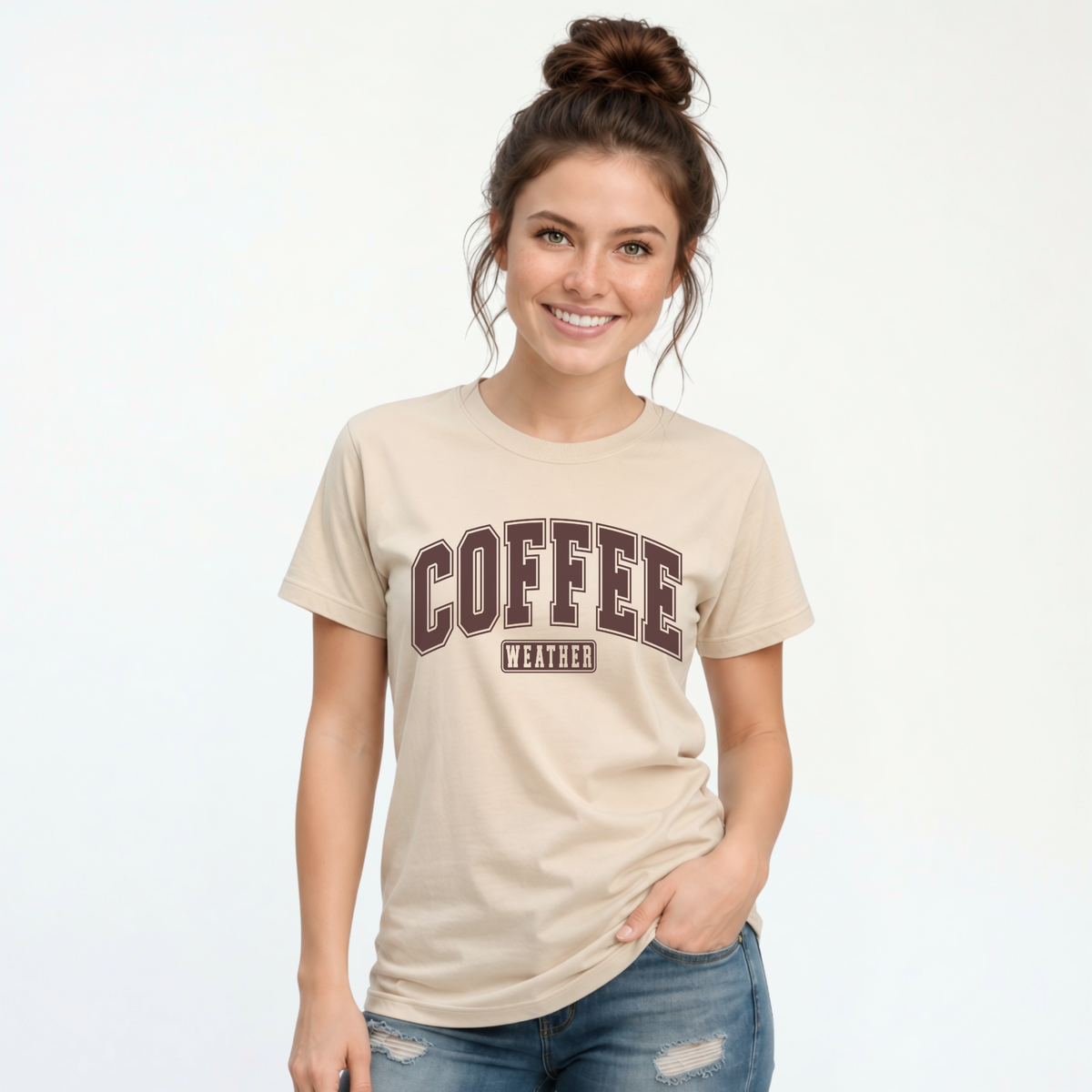 Woman modeling a sand T-shirt with "COFFEE WEATHER" printed in dark brown varsity-style lettering on the front, paired with distressed blue jeans.