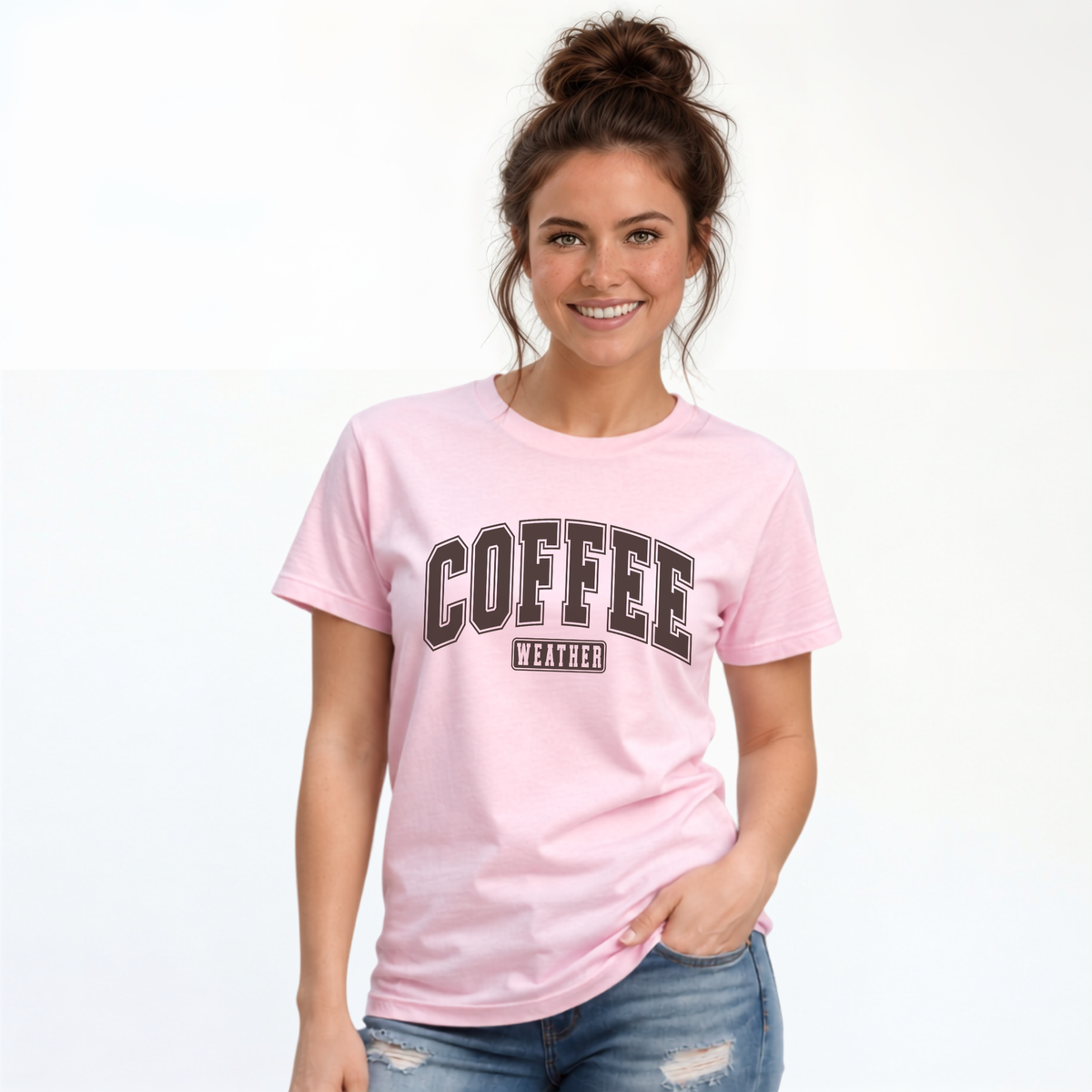 Woman modeling a light pink T-shirt with "COFFEE WEATHER" printed in dark brown varsity-style lettering across the chest, paired with distressed blue jeans.