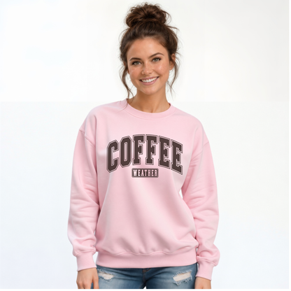 Smiling woman wearing a light pink crewneck sweatshirt with "COFFEE WEATHER" printed in dark brown varsity lettering on the front, paired with distressed blue jeans.