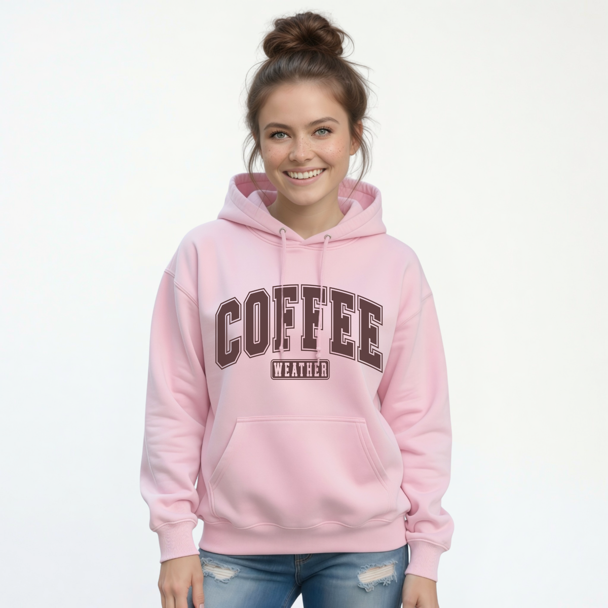 Smiling woman wearing a light pink pullover hoodie with "COFFEE WEATHER" printed in dark brown varsity-style letters on the front, paired with distressed blue jeans.