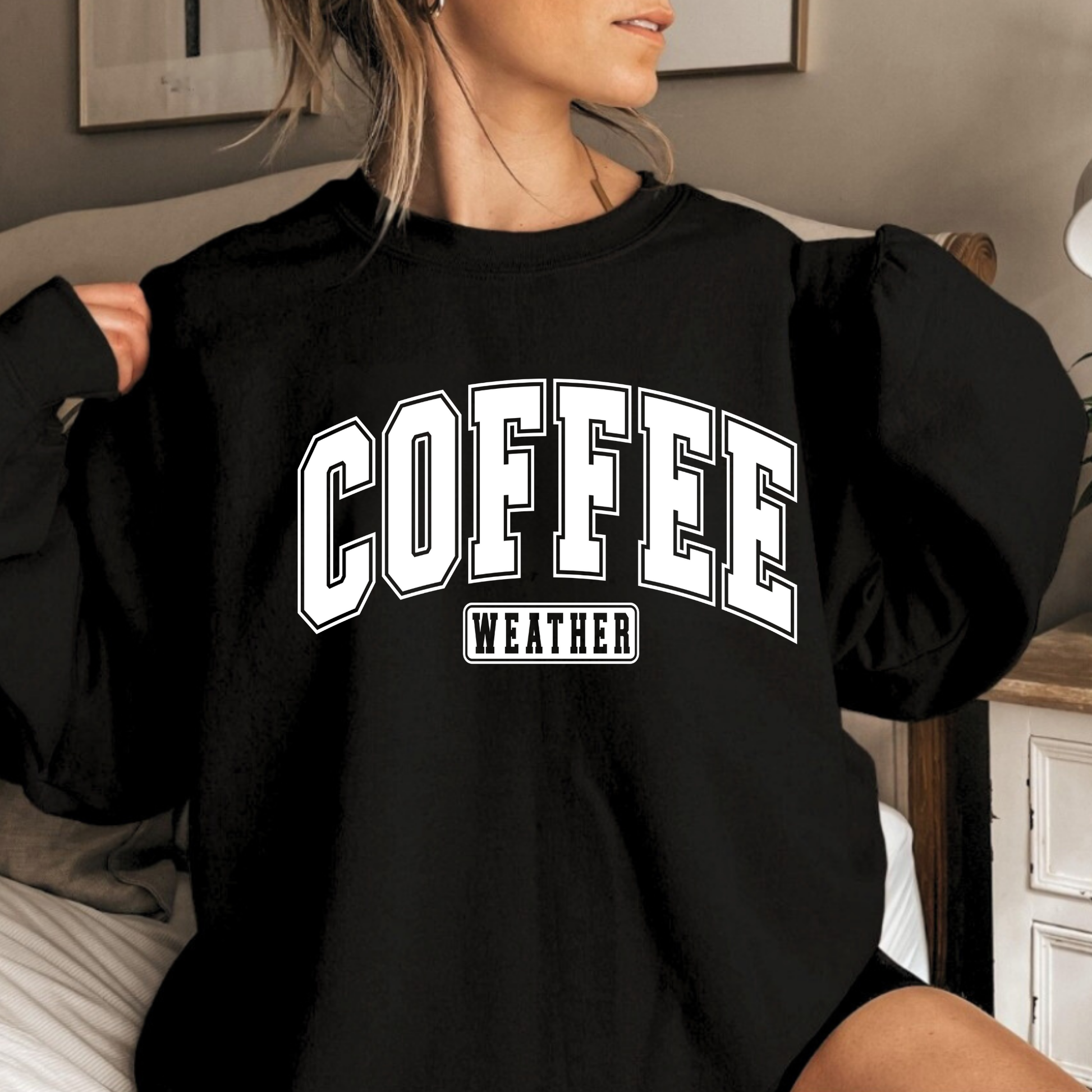 Woman wearing Black "Coffee Weather" sweatshirt sitting cozy on bed

