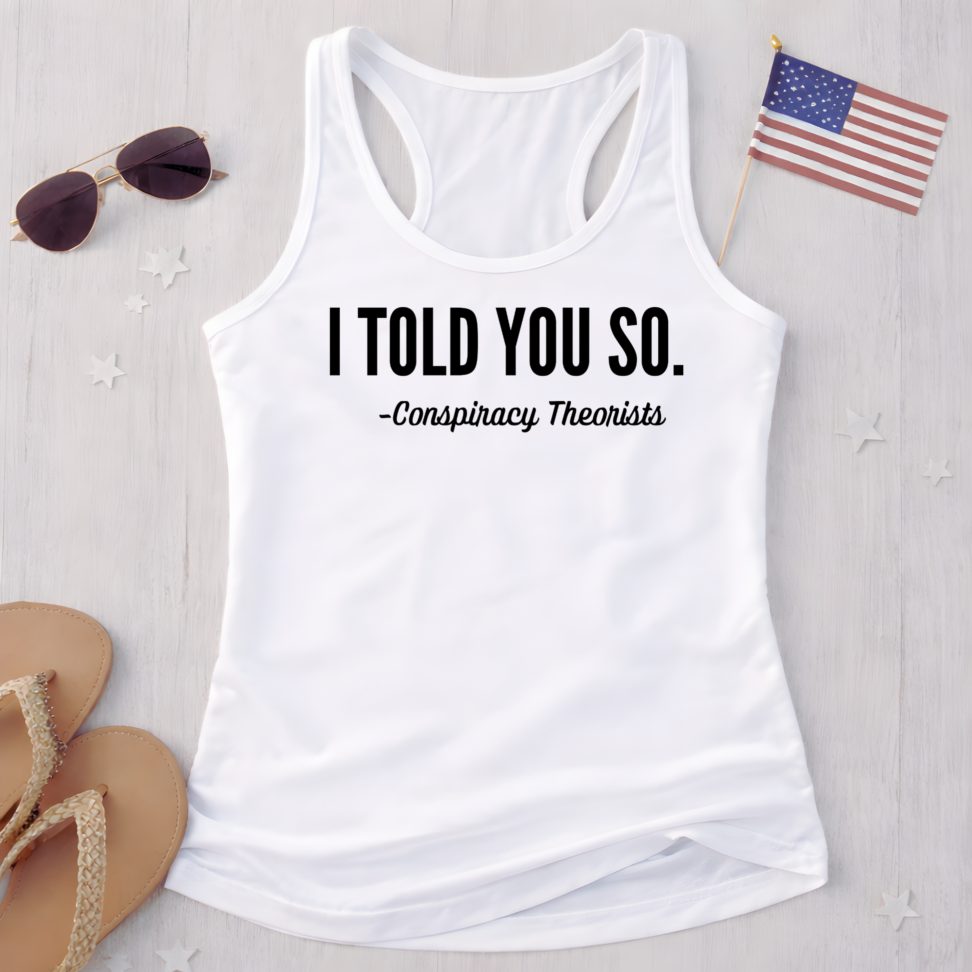 White tank top with text 'I Told You So. - Conspiracy Theorists' on a light wooden surface with sunglasses and an American flag.