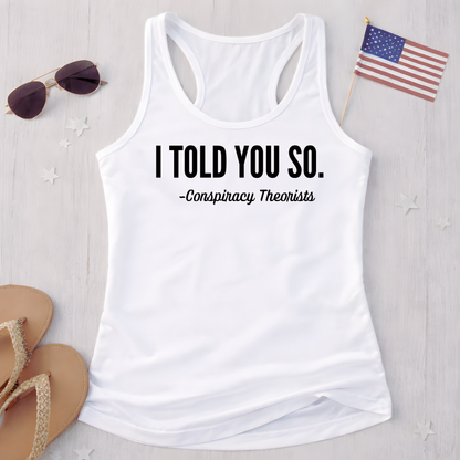 White tank top with text 'I Told You So. - Conspiracy Theorists' on a light wooden surface with sunglasses and an American flag.