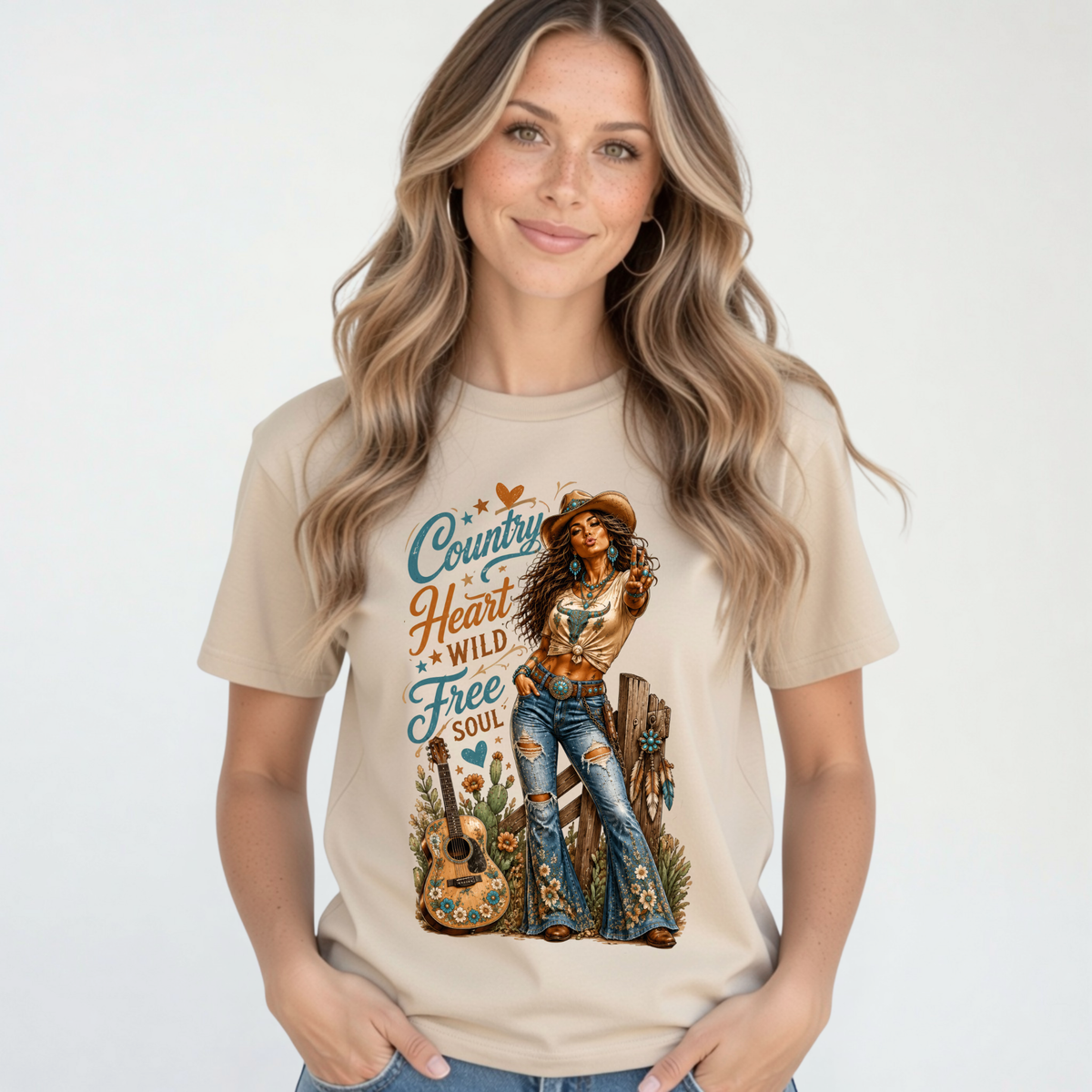 Blonde woman modeling a sand T-shirt featuring the Country Heart Wild Free Soul western cowgirl graphic in a simple studio setting.