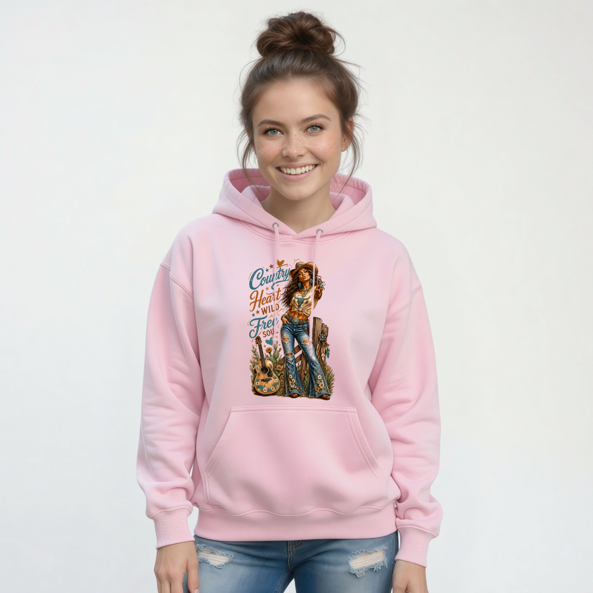 Woman wearing a light pink hoodie printed with the Country Heart Wild Free Soul country cowgirl design against a white studio backdrop.