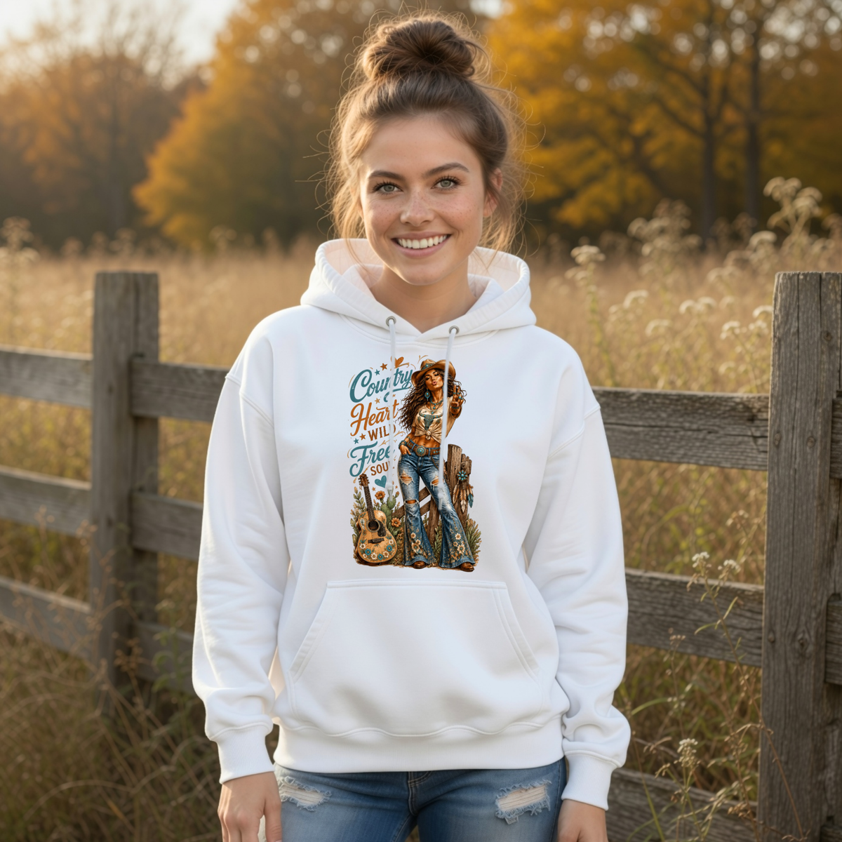 Woman in a white hoodie with the Country Heart Wild Free Soul western cowgirl design beside a rustic fence in an autumn field.