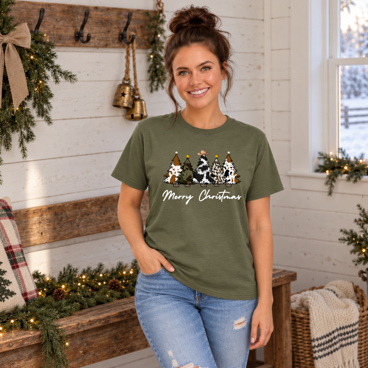 Woman modeling a military green cow print Christmas trees T-shirt beside a festive farmhouse bench.