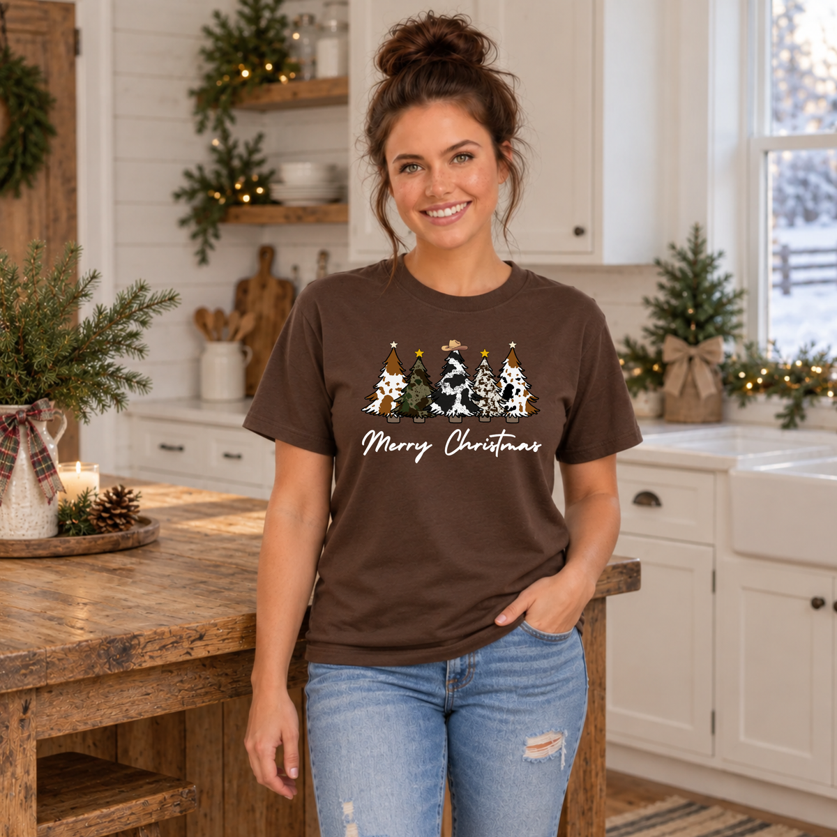 Woman modeling a brown cow print Christmas trees T-shirt beside a farmhouse table.