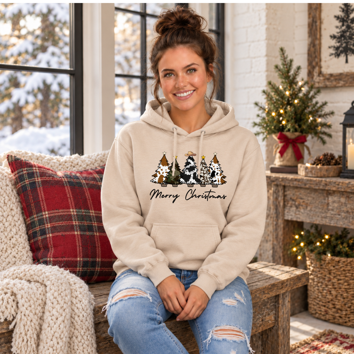 Woman seated in a sand cow print Christmas trees hoodie in a cozy Christmas room.