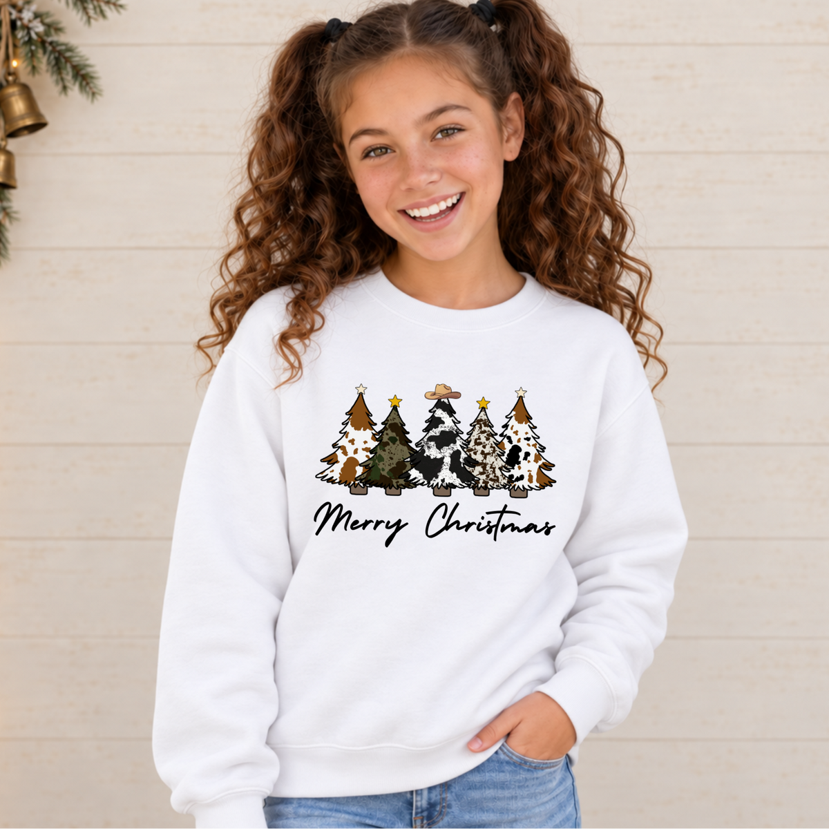 Girl wearing a white Merry Christmas cow print trees sweatshirt against a light shiplap wall with holiday lights.