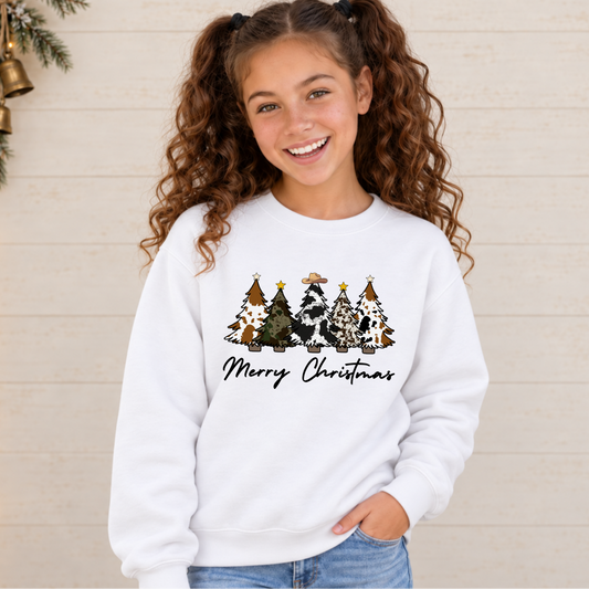 Girl wearing a white Merry Christmas cow print trees sweatshirt against a light shiplap wall with holiday lights.