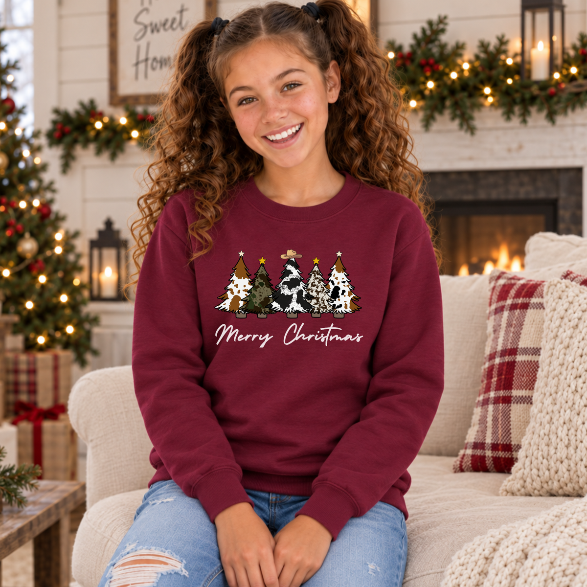 Girl seated in a maroon Merry Christmas cow print trees sweatshirt beside a Christmas tree and fireplace.