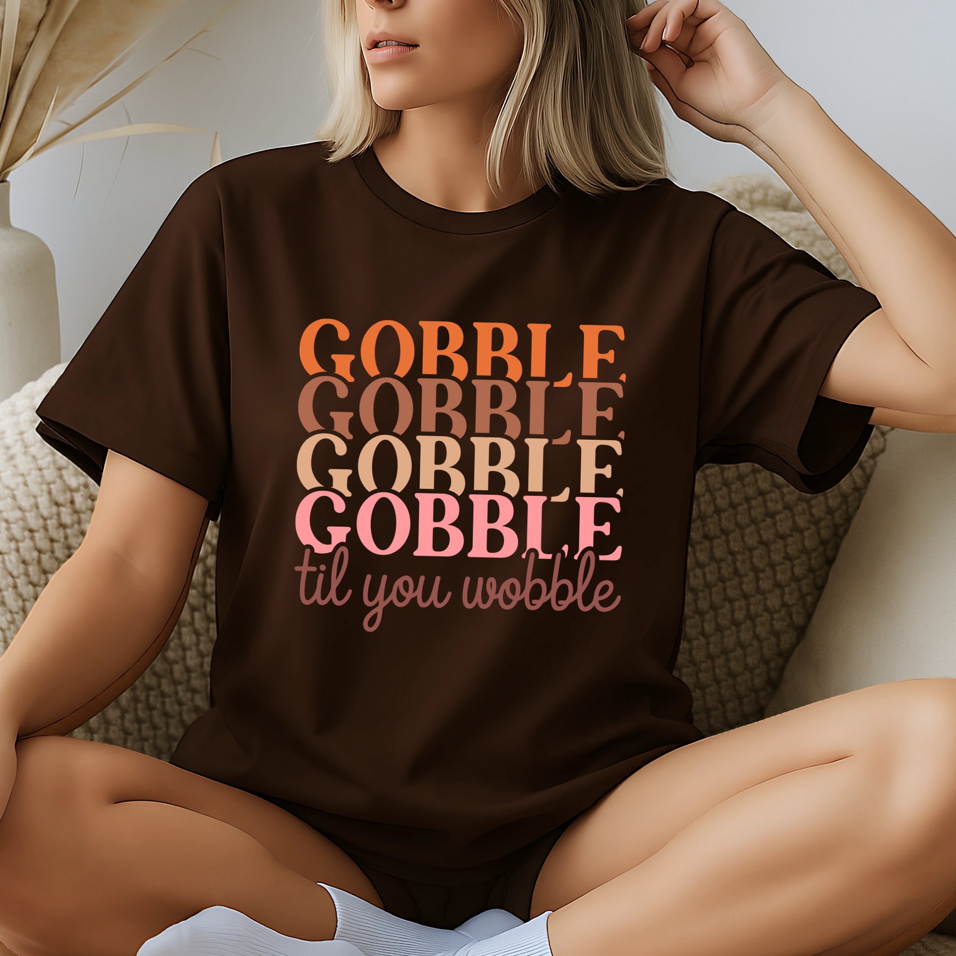 Cozy indoor setting with a woman lounging in a dark brown Thanksgiving t-shirt featuring “Gobble Til You Wobble” in orange and pink tones