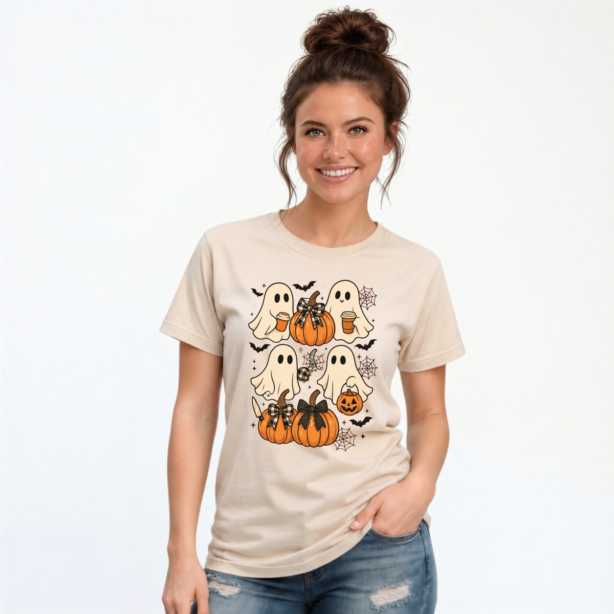 A woman wearing a beige graphic t-shirt with a cute Halloween print of ghosts, pumpkins, and bats, paired with blue jeans, against a plain white background.