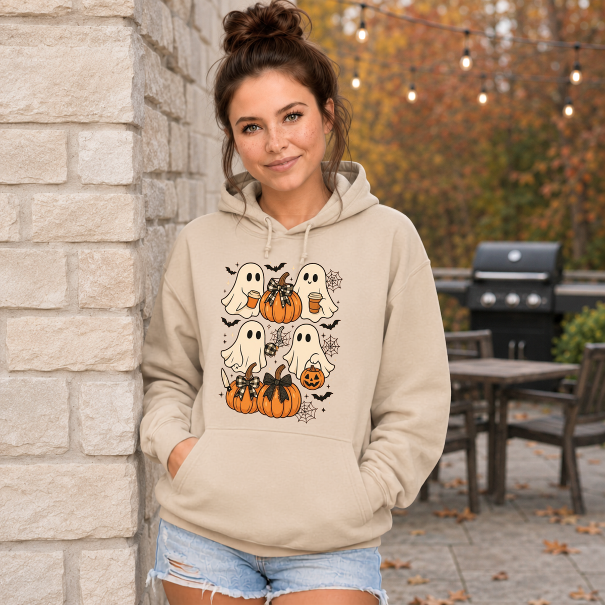 A woman wearing a beige pullover hoodie with a Halloween graphic print featuring cartoon ghosts, pumpkins, bats, and spiderwebs, paired with blue jeans against a plain white background.