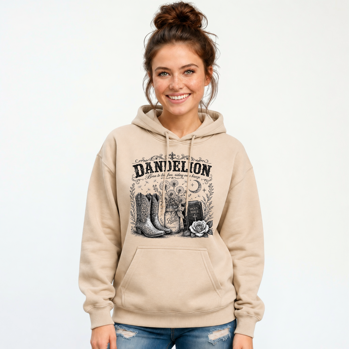 Woman wearing a sand Dandelion pullover hoodie with cowboy boots, dandelion-filled mason jar, Holy Bible, rose, and crescent moon graphic on a white background