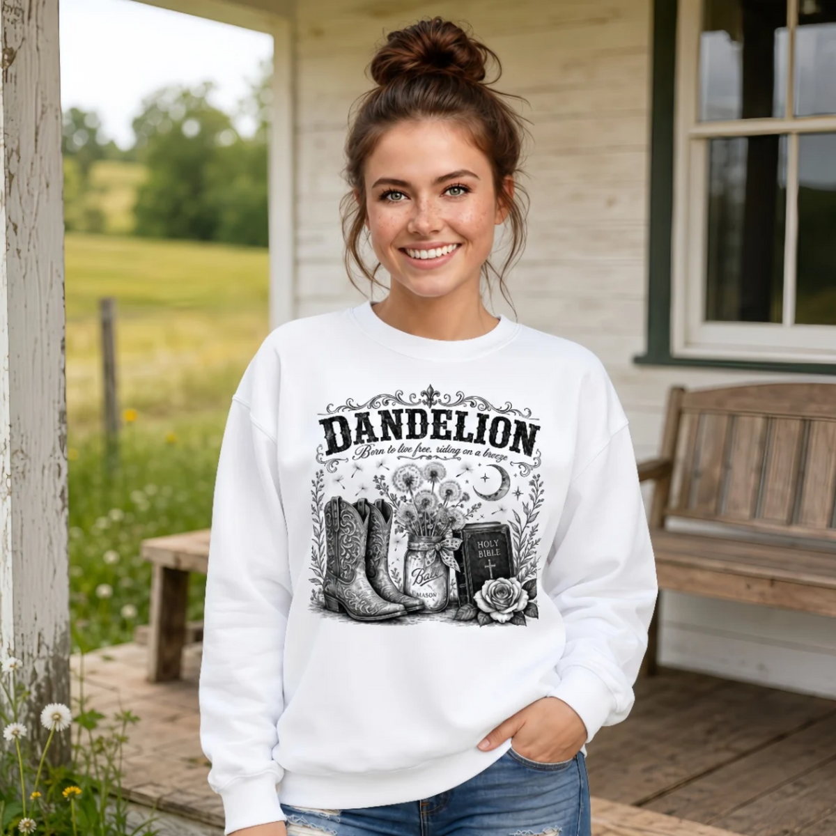 Woman wearing a white Dandelion crewneck sweatshirt with cowboy boots, dandelion-filled mason jar, Holy Bible, rose, and crescent moon graphic on a farmhouse porch