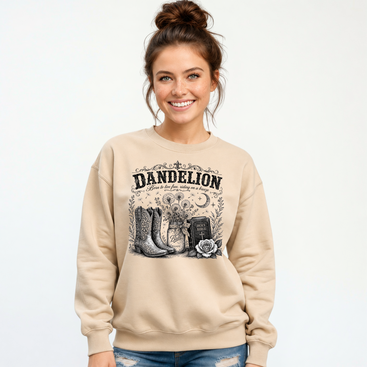Woman wearing a sand Dandelion crewneck sweatshirt with cowboy boots, dandelion-filled mason jar, Holy Bible, rose, and crescent moon graphic on a white background