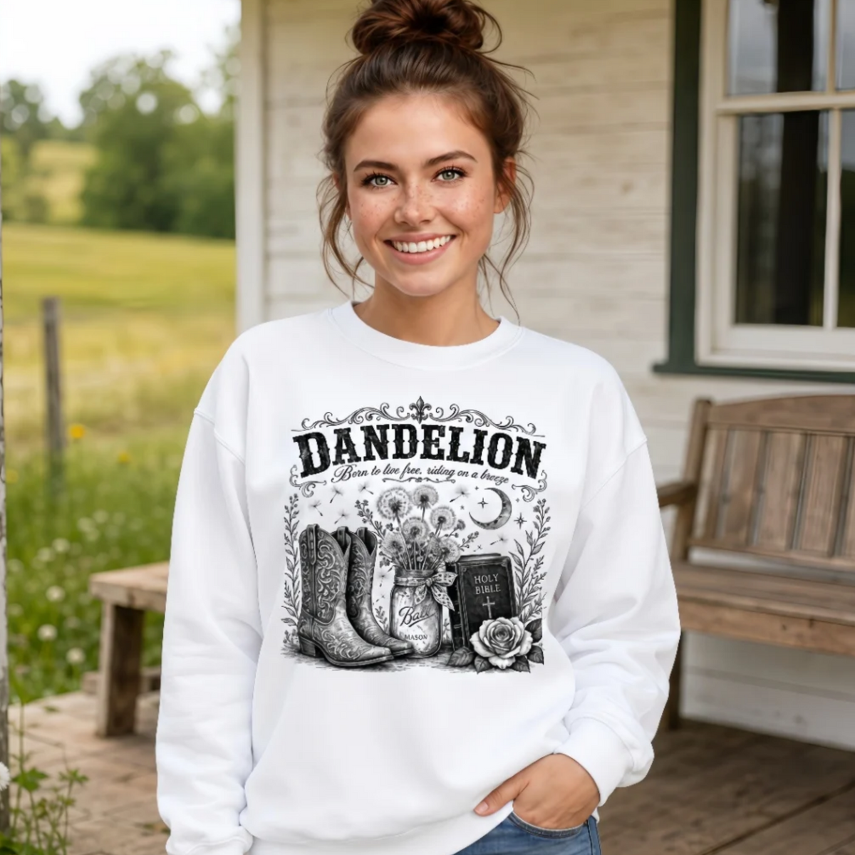 Woman wearing a white Dandelion crewneck sweatshirt with cowboy boots, dandelion-filled mason jar, Holy Bible, rose, and crescent moon graphic on a farmhouse porch