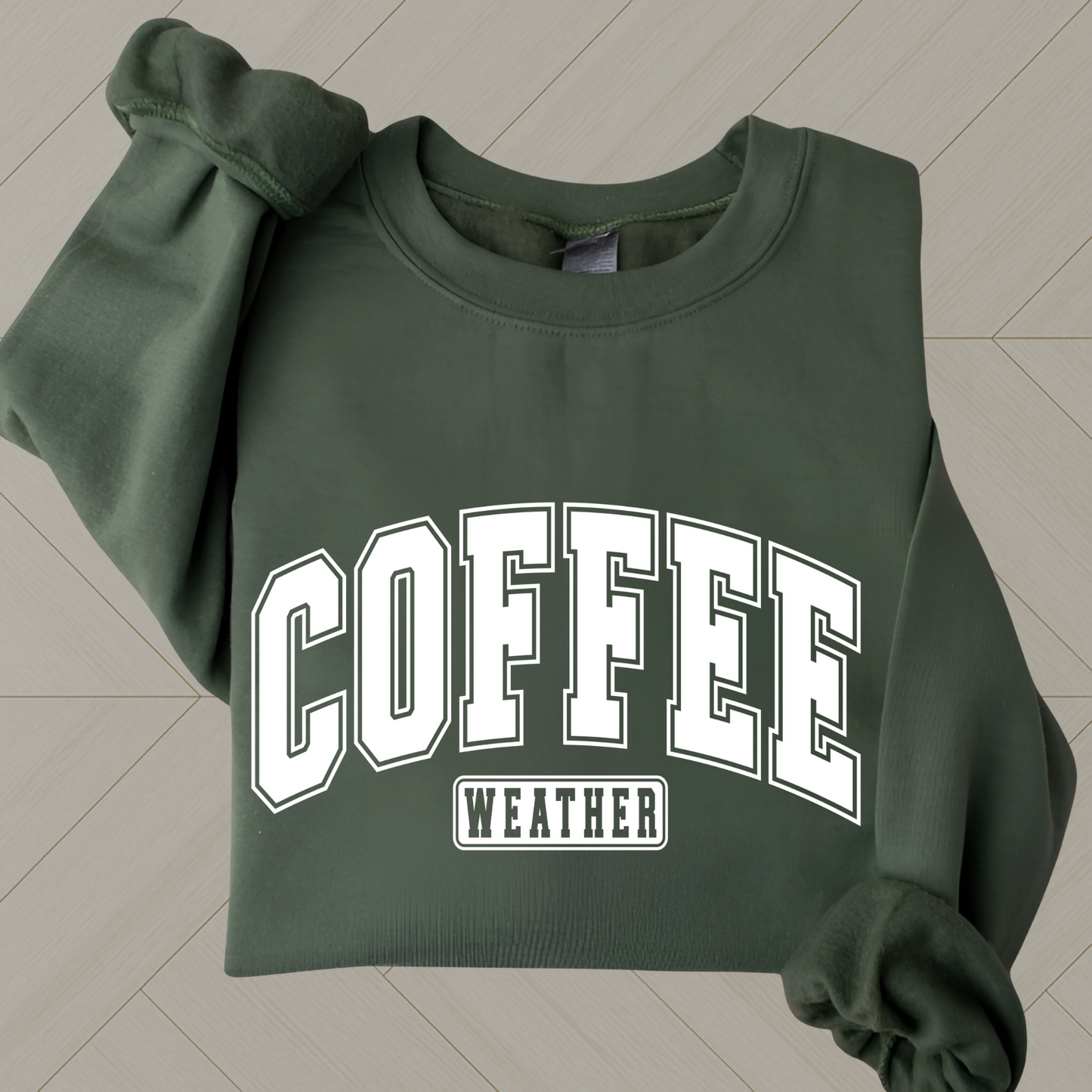 Dark green folded sweatshirt with bold white “Coffee Weather” text – flat lay on wood floor


