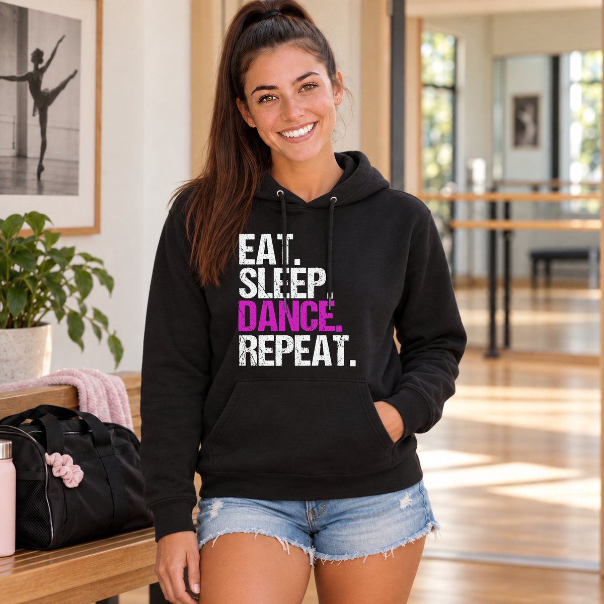 Woman wearing a black Eat Sleep Dance Repeat hoodie in a dance studio lobby beside a dance bag.