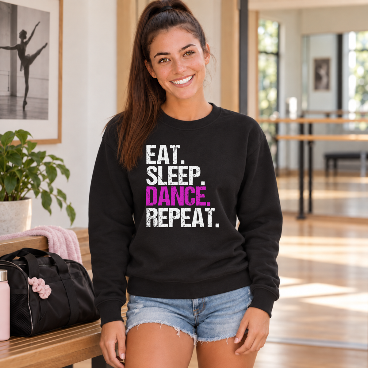 Woman in a black Eat Sleep Dance Repeat sweatshirt beside a bench and dance bag in a studio hallway.