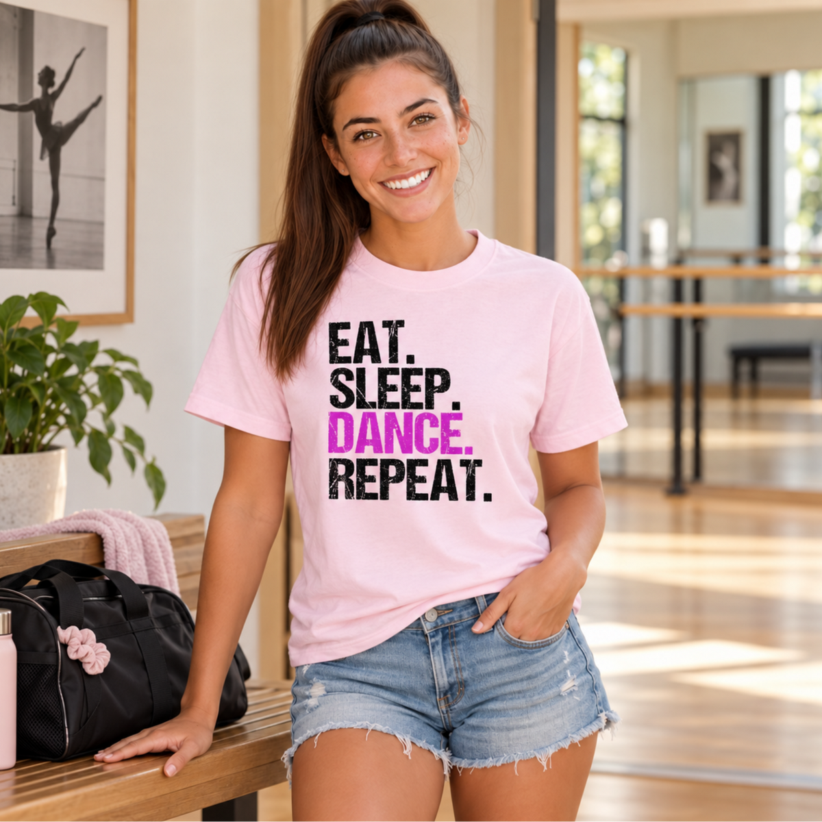Woman wearing a light pink Eat Sleep Dance Repeat T-shirt beside a dance bag in a studio lobby.