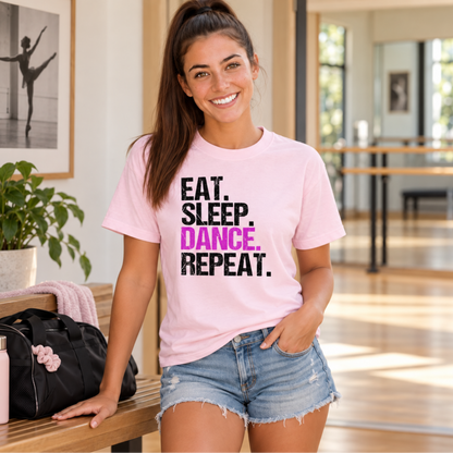 Woman wearing a light pink Eat Sleep Dance Repeat T-shirt beside a dance bag in a studio lobby.