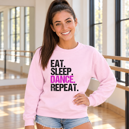 Woman in a light pink Eat Sleep Dance Repeat sweatshirt standing by the barre in a sunlit dance studio.