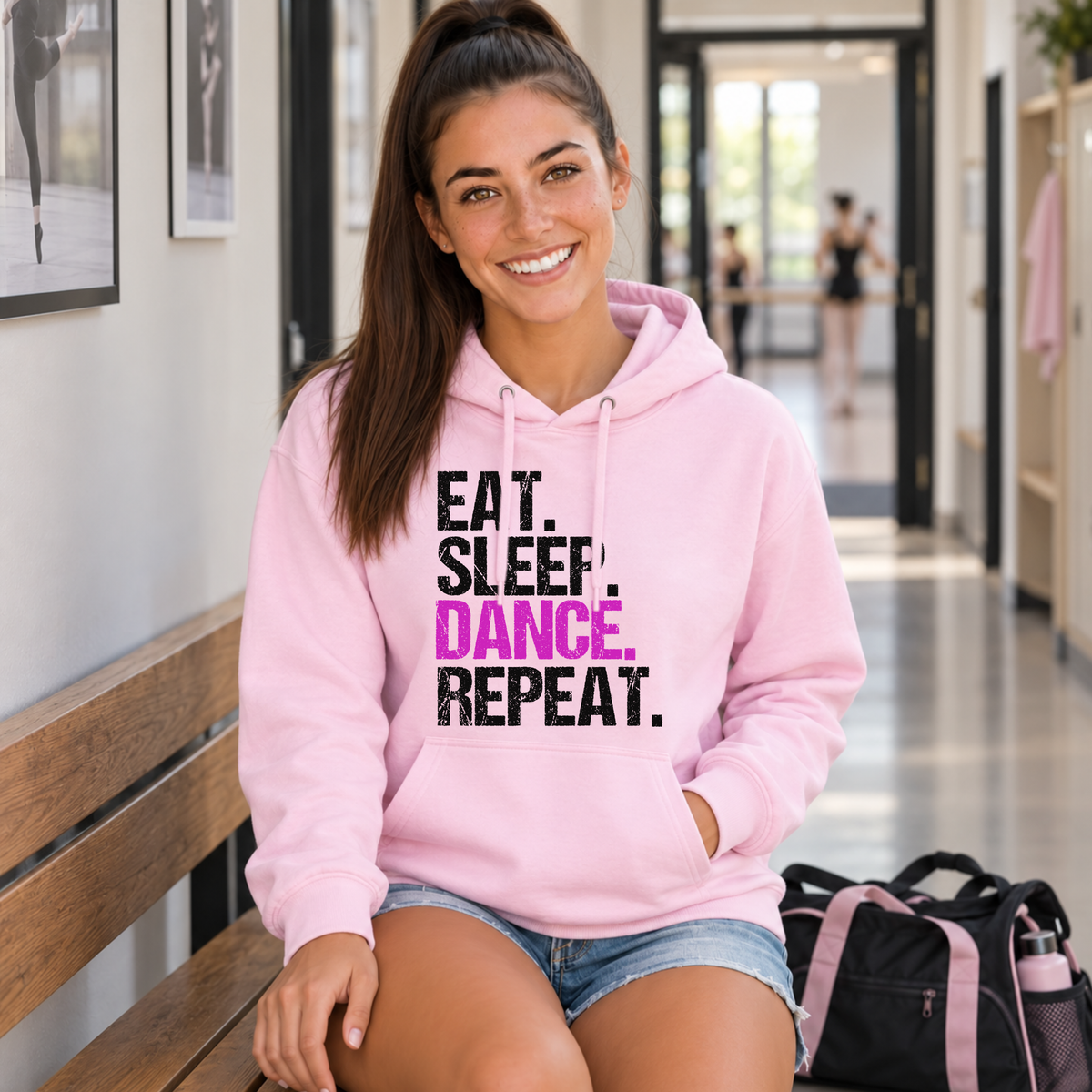Woman seated on a dance studio bench in a light pink Eat Sleep Dance Repeat hoodie beside her dance bag.