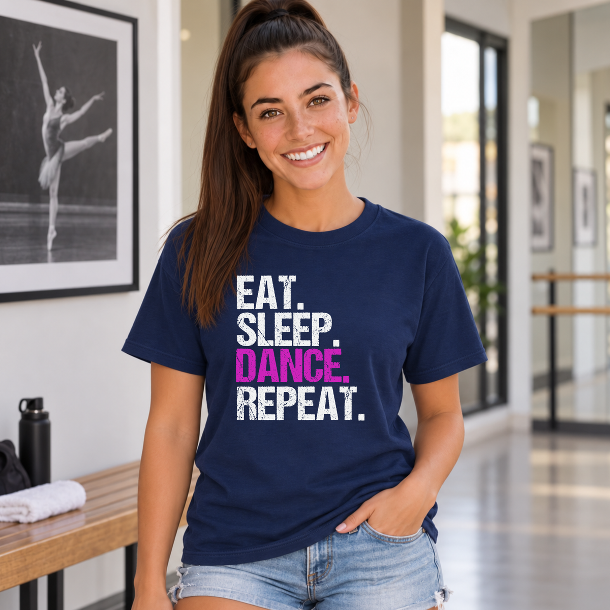 Woman wearing a navy Eat Sleep Dance Repeat T-shirt near a dance studio bench and dancer portrait.