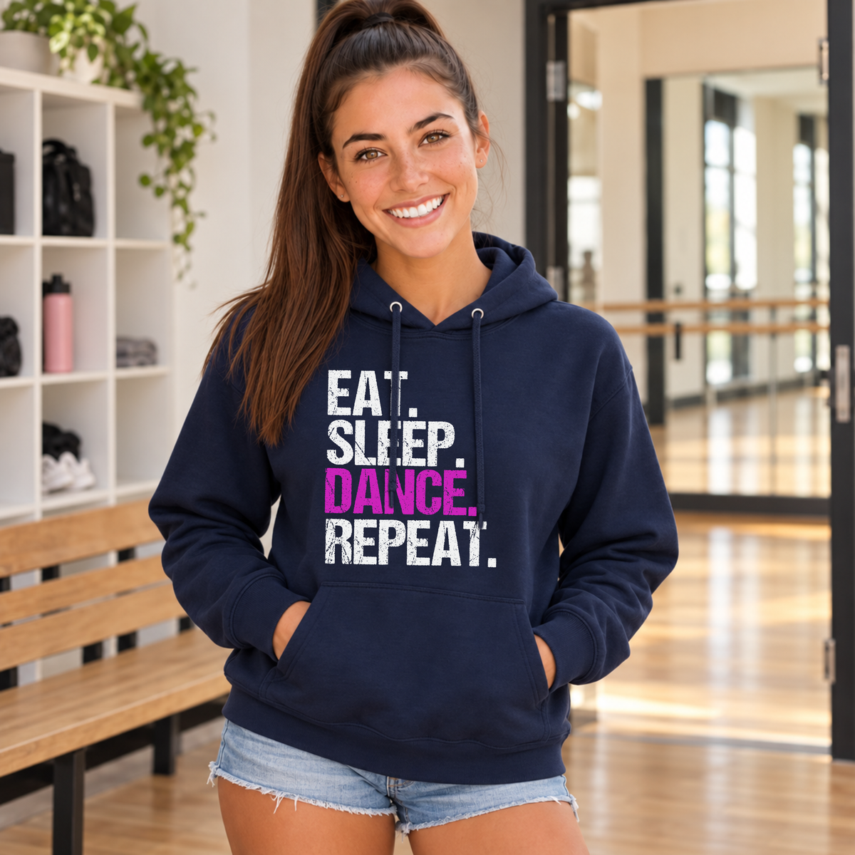 Woman in a navy Eat Sleep Dance Repeat hoodie in a dance studio lobby with cubby shelves.