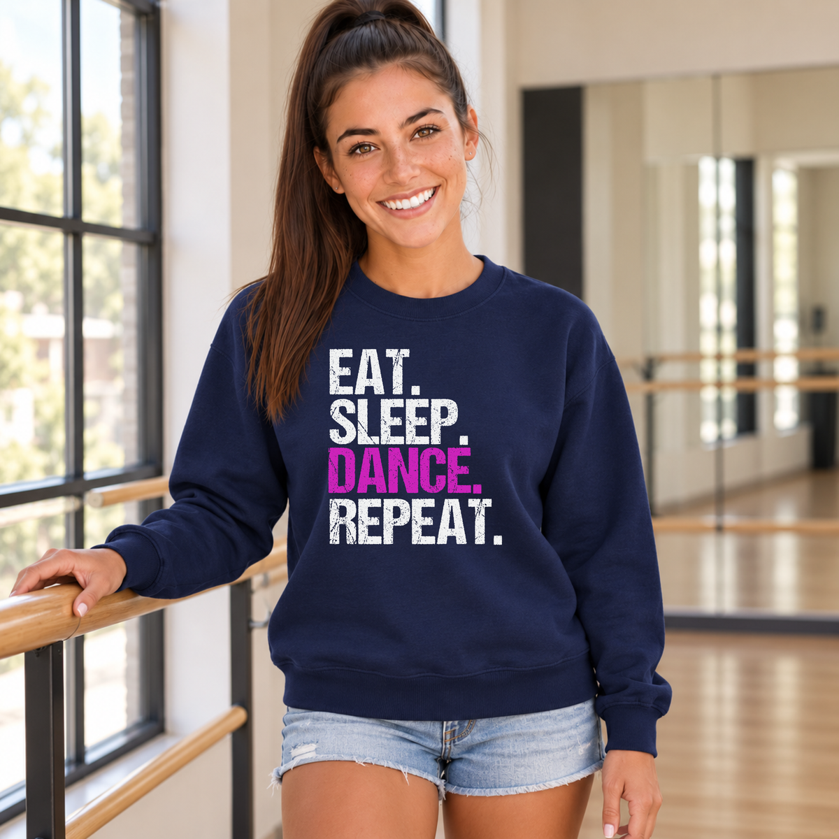 Woman in a navy Eat Sleep Dance Repeat sweatshirt beside a barre and windows in a dance studio.
