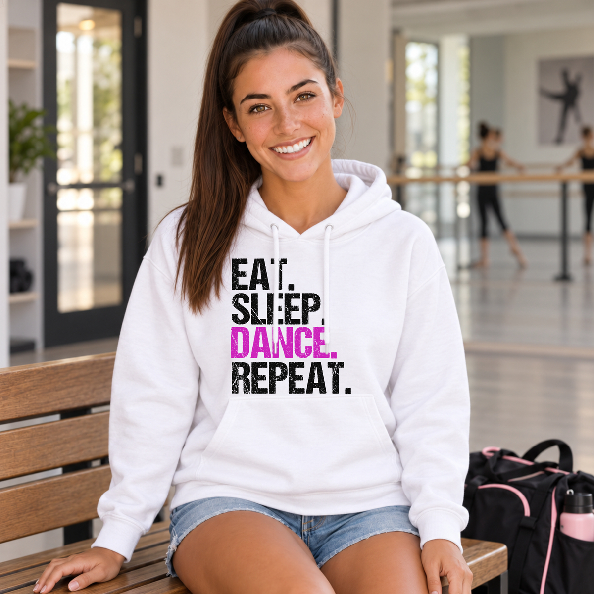 Woman seated on a studio bench in a white Eat Sleep Dance Repeat hoodie beside a dance bag.