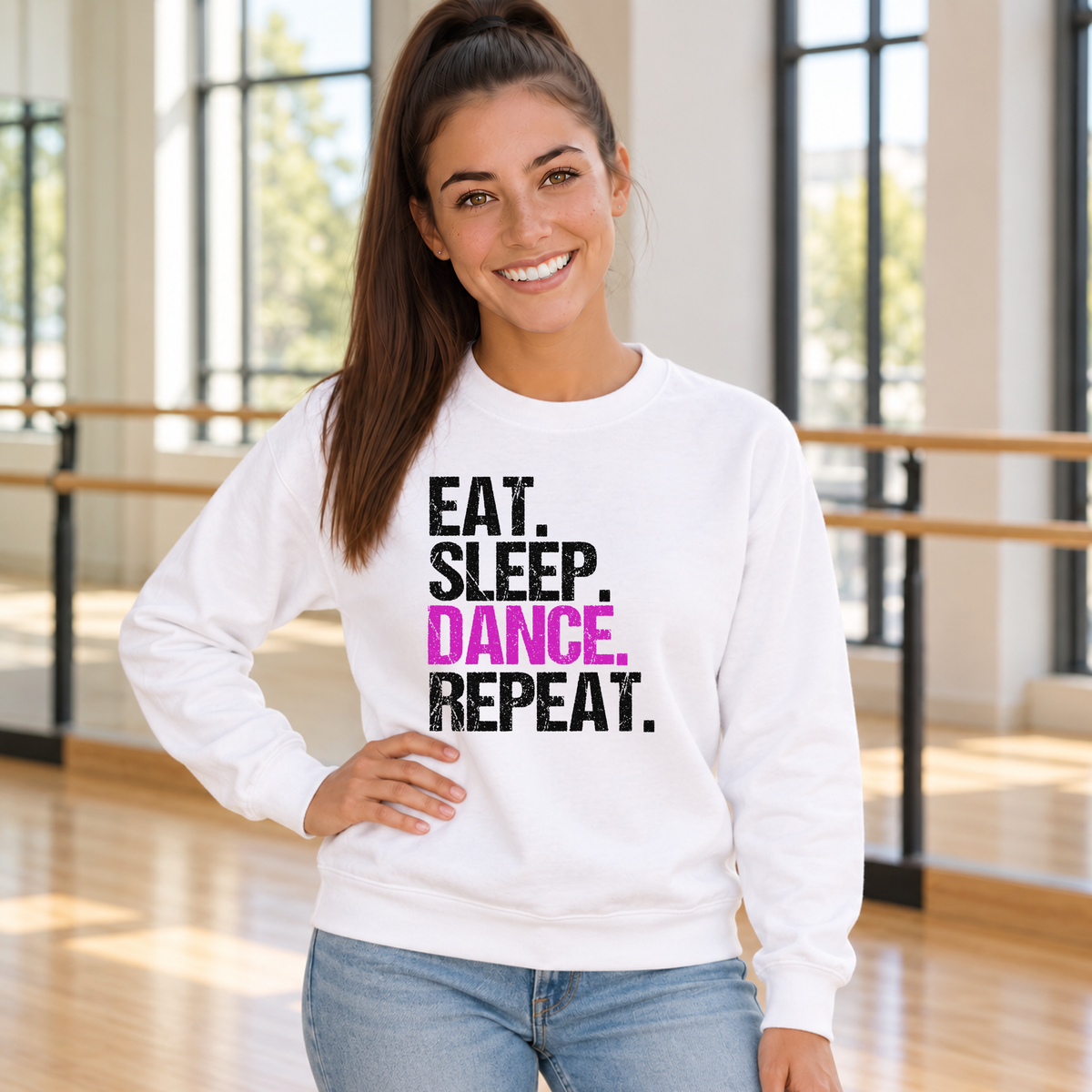 Woman wearing a white Eat Sleep Dance Repeat sweatshirt in a sunlit studio with ballet barres.