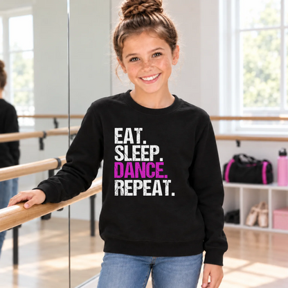 Girl wearing a black youth Eat Sleep Dance Repeat sweatshirt beside a ballet barre and dance bag.
