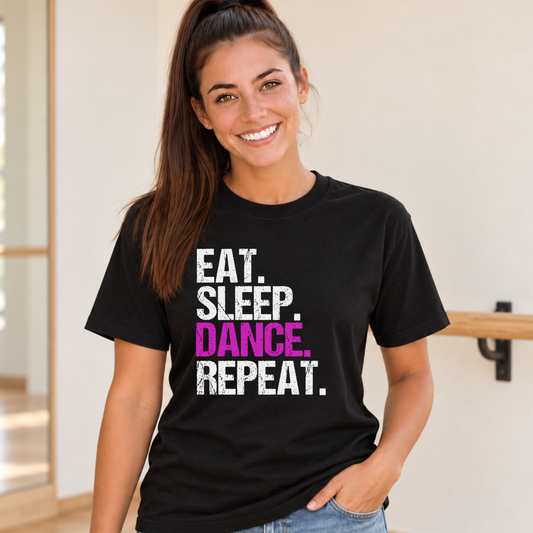 Woman wearing a black Eat Sleep Dance Repeat T-shirt with pink DANCE lettering against a light dance studio wall.