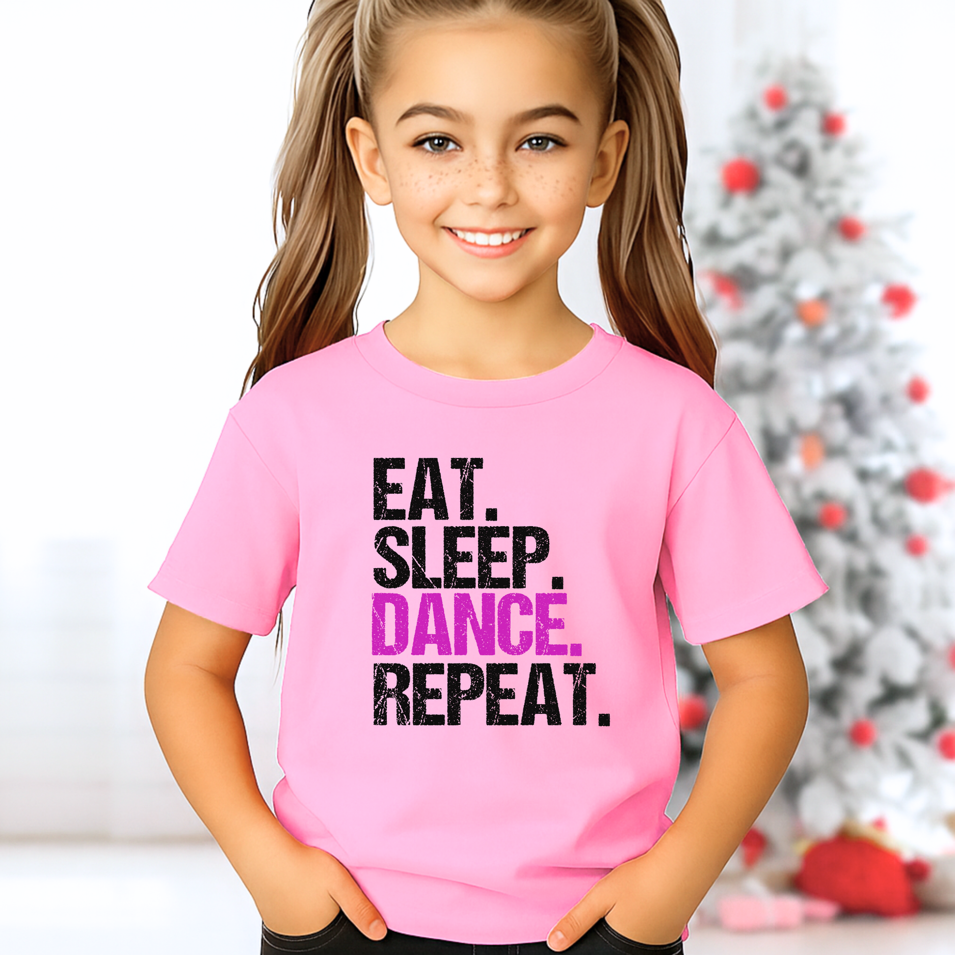 Girl in light pink youth dance t-shirt standing in Christmas-themed background, “Eat Sleep Dance Repeat” graphic, cute holiday dancer gift.
