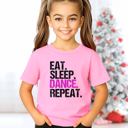 Girl in light pink youth dance t-shirt standing in Christmas-themed background, “Eat Sleep Dance Repeat” graphic, cute holiday dancer gift.