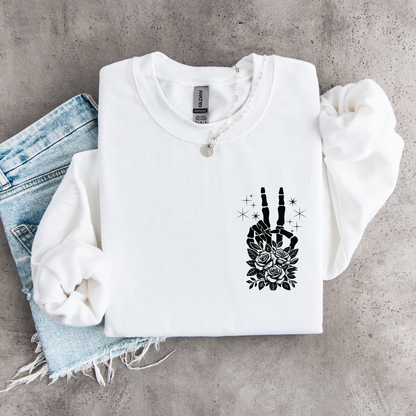 White crewneck sweatshirt flat lay with a small black floral skeleton peace-sign graphic on the left chest, styled with denim.