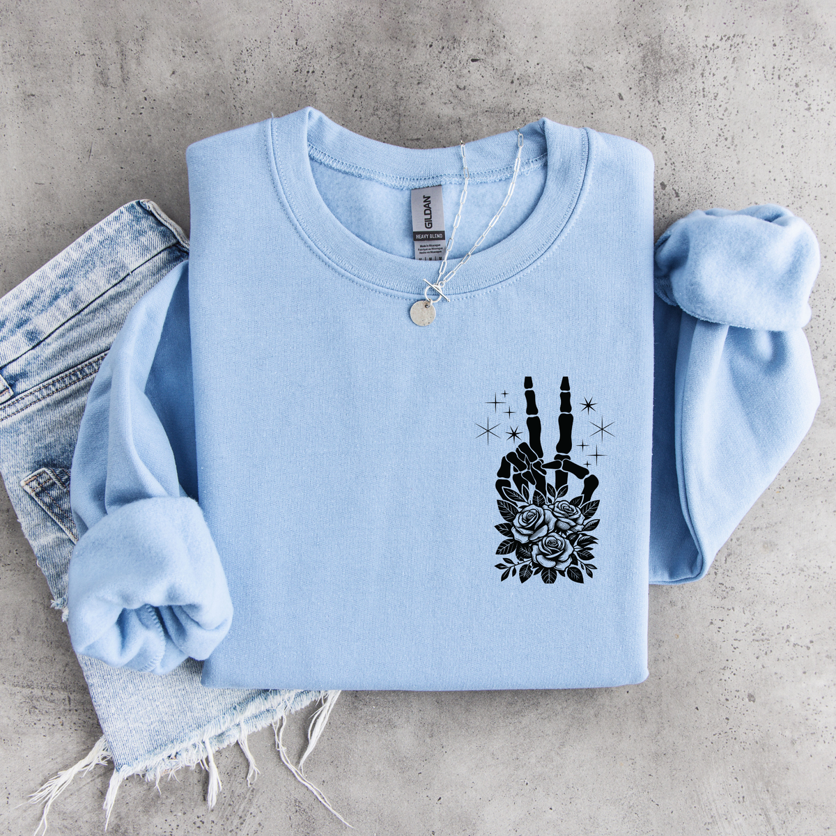 Light blue crewneck sweatshirt flat lay with a small black floral skeleton peace-sign graphic on the left chest, styled with denim.