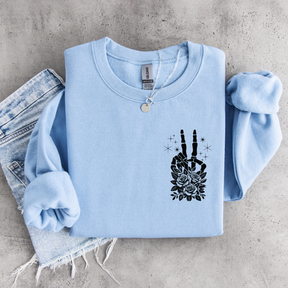 Light blue crewneck sweatshirt flat lay with a small black floral skeleton peace-sign graphic on the left chest, styled with denim.