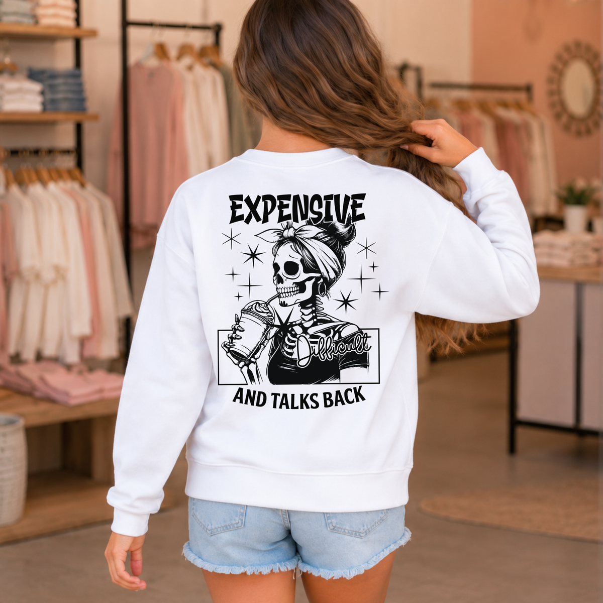Young girl showing the back of a white crewneck sweatshirt with a black Expensive and Talks Back skeleton-woman graphic in a boutique setting.