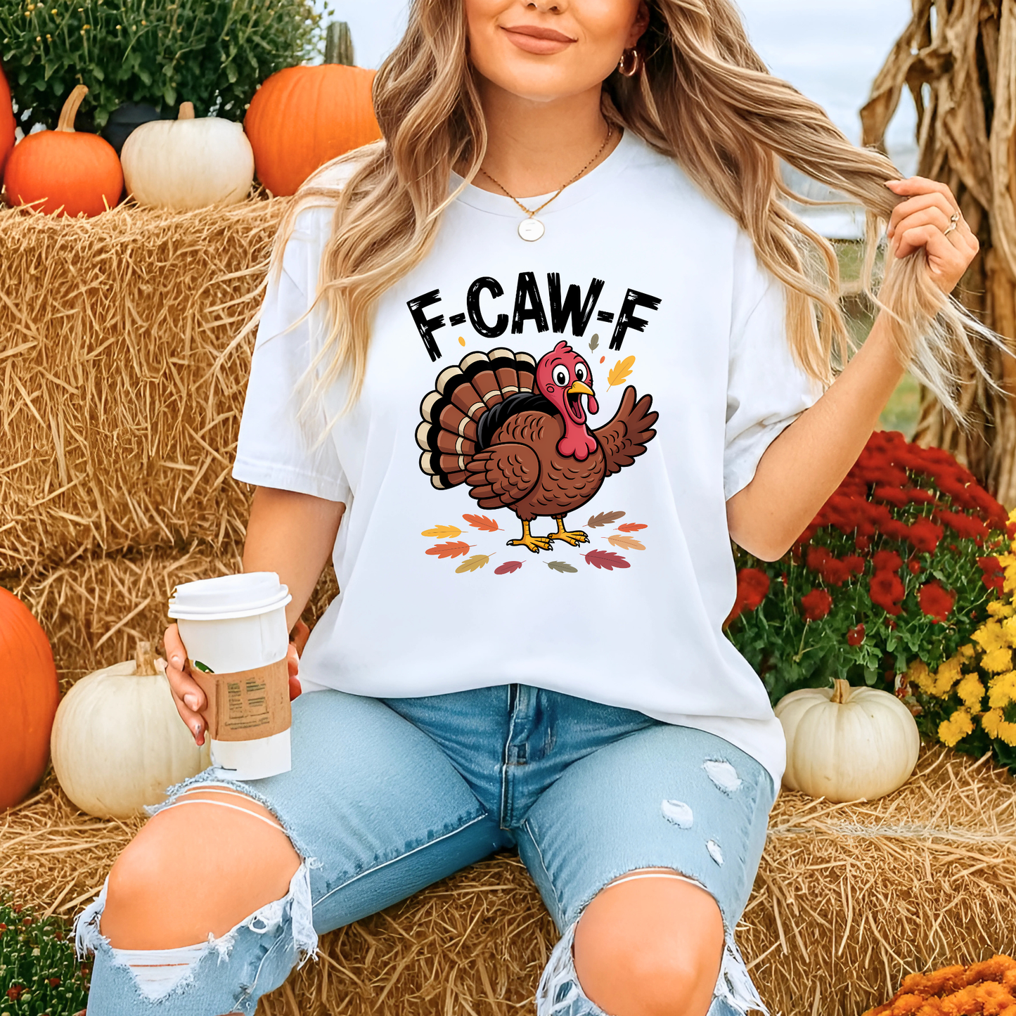 Woman sitting outdoors at a fall pumpkin patch wearing white “F-CAW-F” turkey shirt and holding coffee
