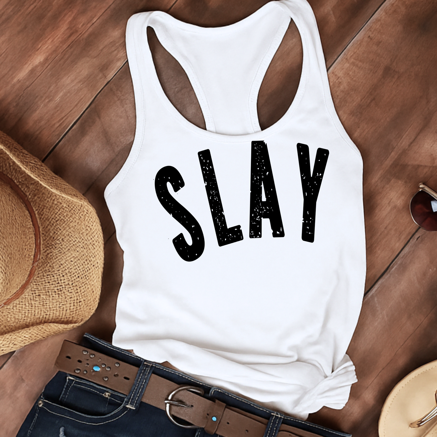 Flat lay of a white racerback tank top featuring large distressed black “SLAY” text, styled with a brown wide-brimmed straw hat, black belt with silver accents, and dark denim jeans. Set against a rustic wooden background, this laid-back aesthetic highlights the tank’s casual summer vibe, perfect for country festivals, vacation wear, or weekend outfits.

