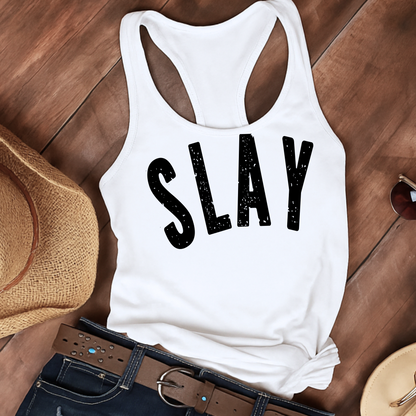 Flat lay of a white racerback tank top featuring large distressed black “SLAY” text, styled with a brown wide-brimmed straw hat, black belt with silver accents, and dark denim jeans. Set against a rustic wooden background, this laid-back aesthetic highlights the tank’s casual summer vibe, perfect for country festivals, vacation wear, or weekend outfits.

